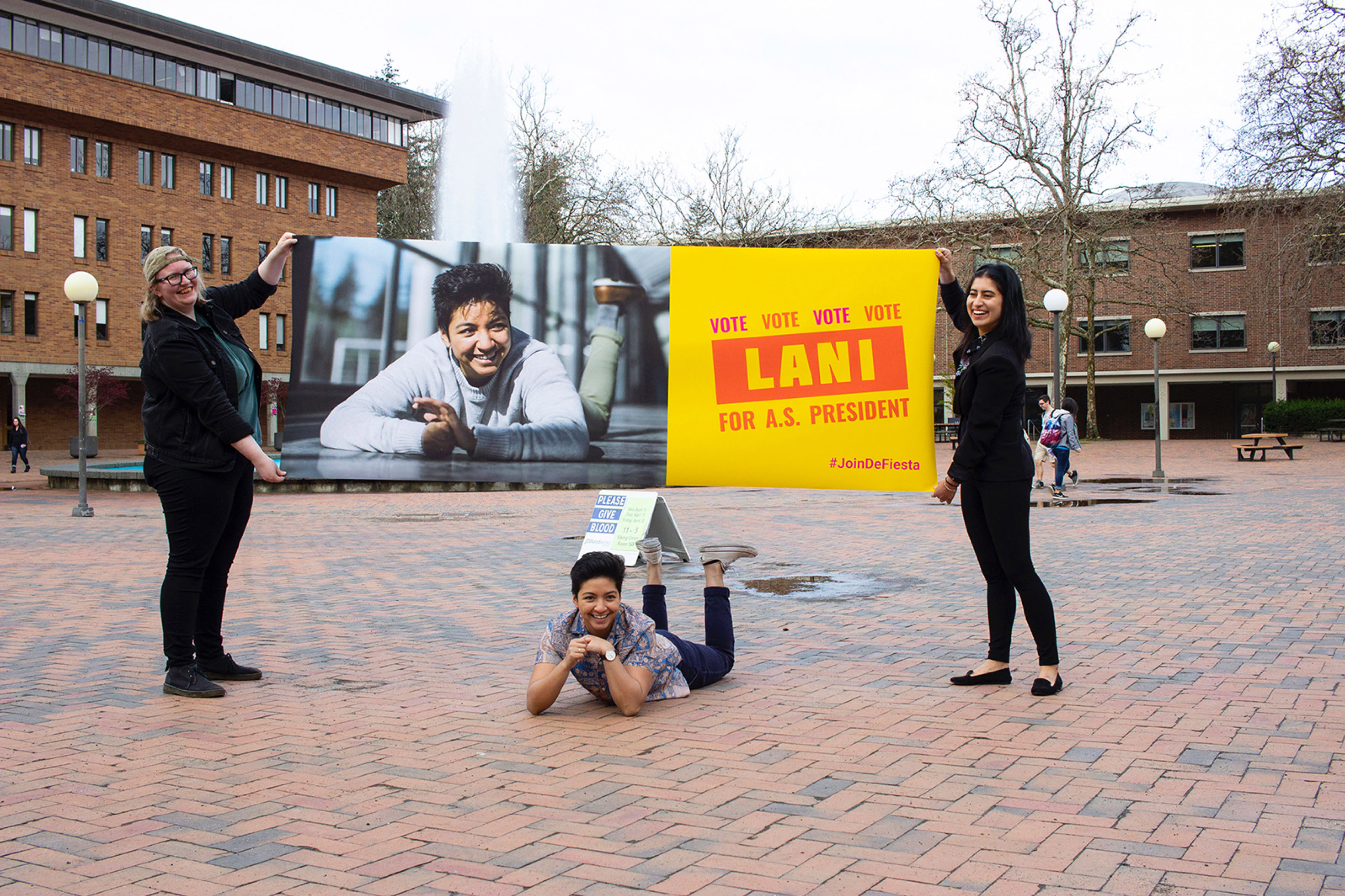 Lani poses under their campaign banner while two members of their campaign team hold it up above them.