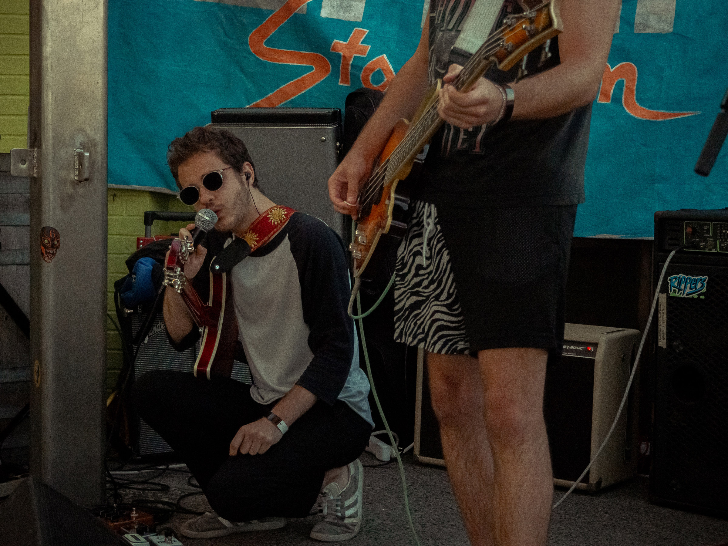 Guitarist, Trevor, kneels over his pedal board with a mic to his mouth. Bassist, Ethan's, legs can be seen in the foreground