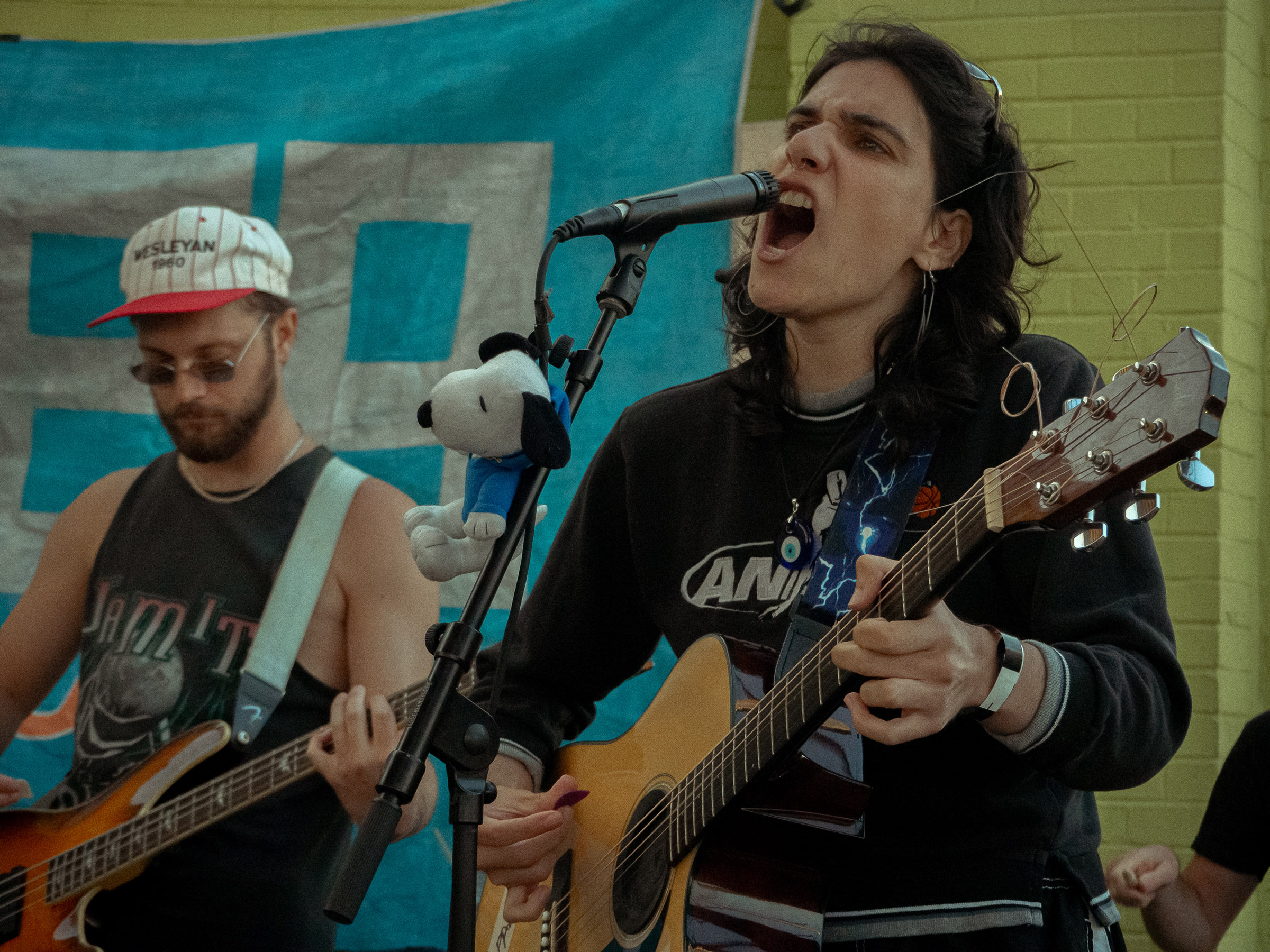 Nara sings aggressively while playing guitar. Bassist, Ethan, can be seen in the background. Nara has a plush Snoopy strapped to their mic stand.